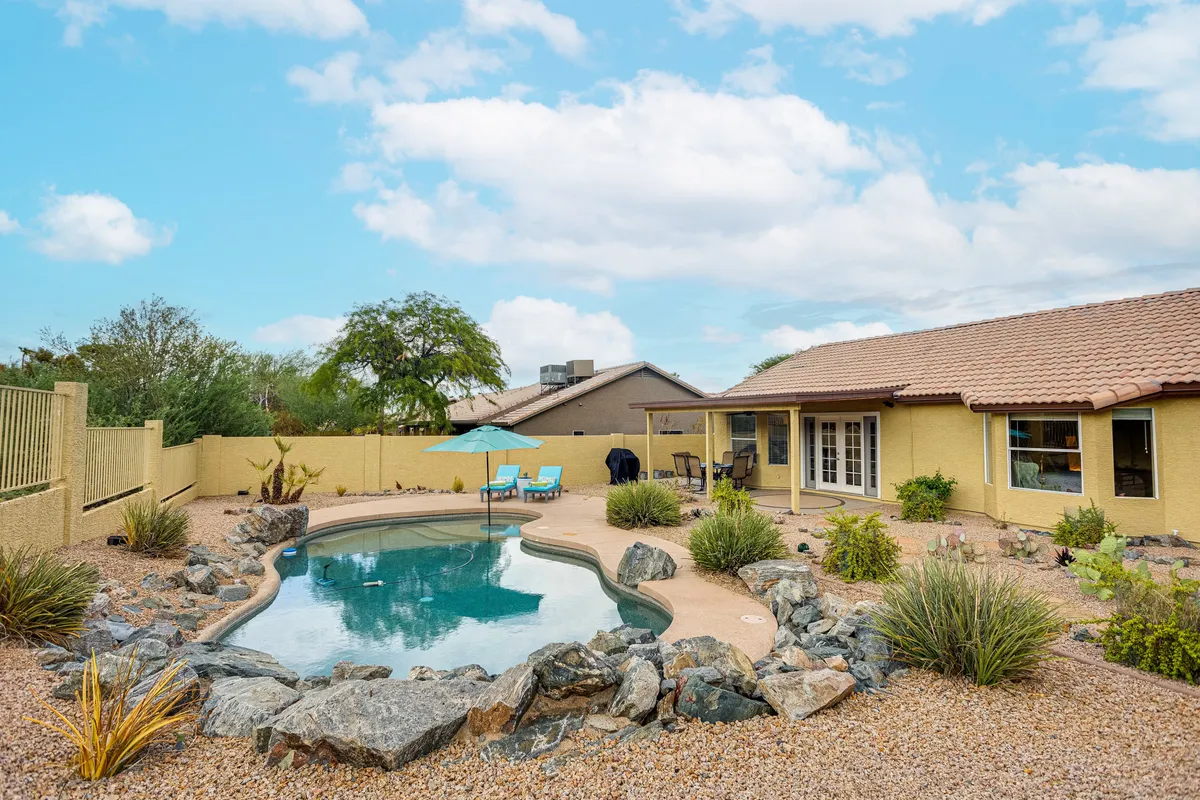 A tranquil backyard scene featuring a sparkling swimming pool surrounded by rock landscaping, with lounge chairs and an inviting patio area shaded by a large umbrella. The space is nestled against natural greenery, offering peaceful views and a relaxing atmosphere.