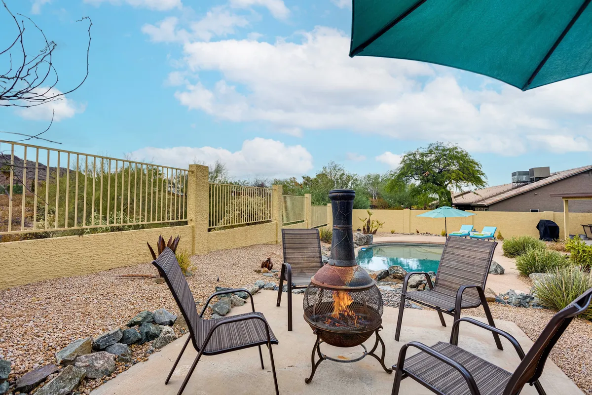An inviting backyard with a fire pit, comfortable outdoor seating under an umbrella, and a clear view of the swimming pool. The space is designed for evening gatherings, relaxation, and enjoying the warmth of the fire under the open sky.
