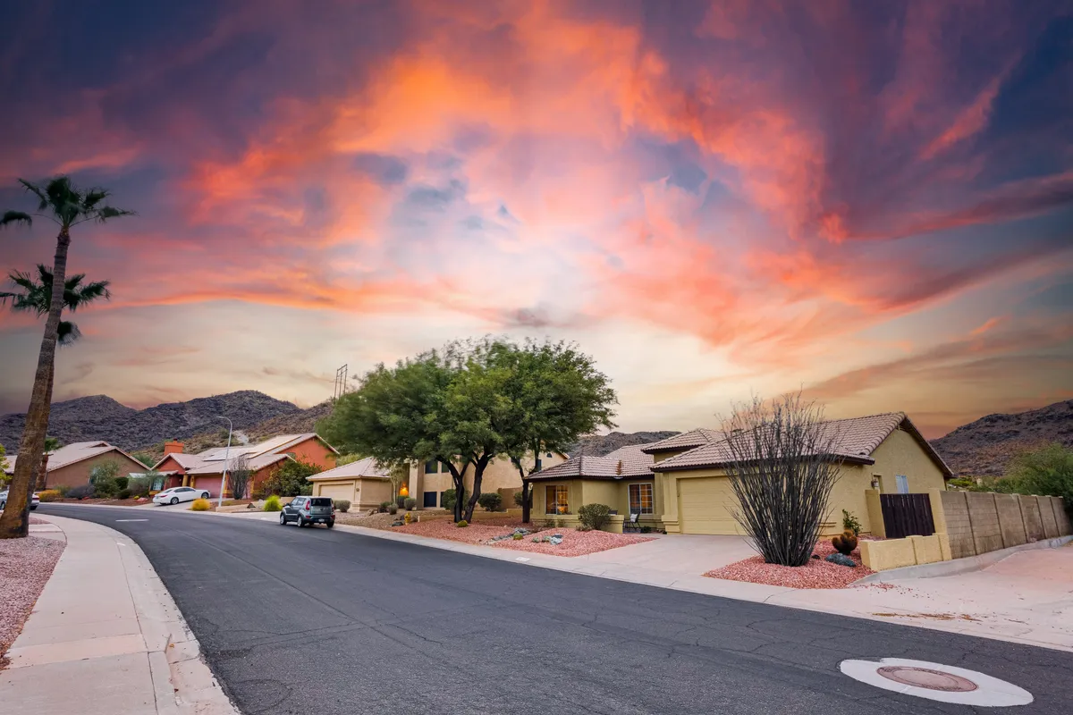 A quiet residential street lined with single-story homes and palm trees, bathed in the vibrant colors of a desert sunset. The sky is ablaze with orange and pink hues, evoking the tranquil beauty of the Arizona landscape and the neighborhood's inviting atmosphere.