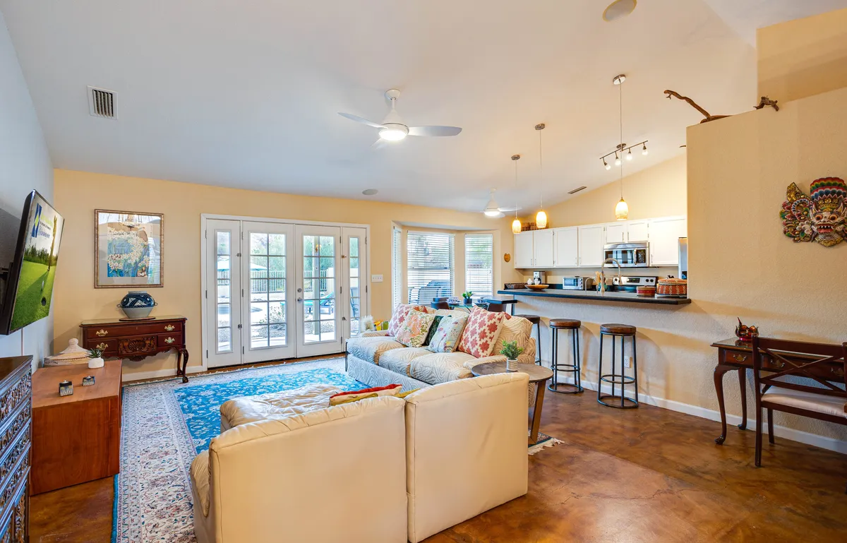 A cheerful, welcoming great room featuring a comfortable sofa with colorful throw pillows, the kitchen bar area, French doors, and artwork on the wall. The open layout visually connects the living, dining, and kitchen spaces, with ample natural light streaming through the doors.