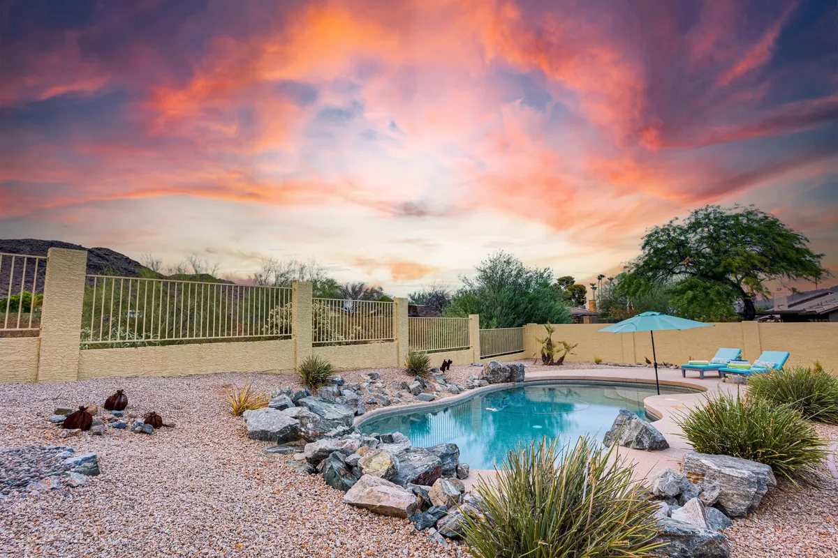 A tranquil backyard scene featuring a sparkling swimming pool surrounded by natural rock formations, lounge chairs, a fence for privacy, and a stunning sunset sky over distant mountains. The setting radiates peace, with ample space for relaxing, sunbathing, or entertaining outdoors.