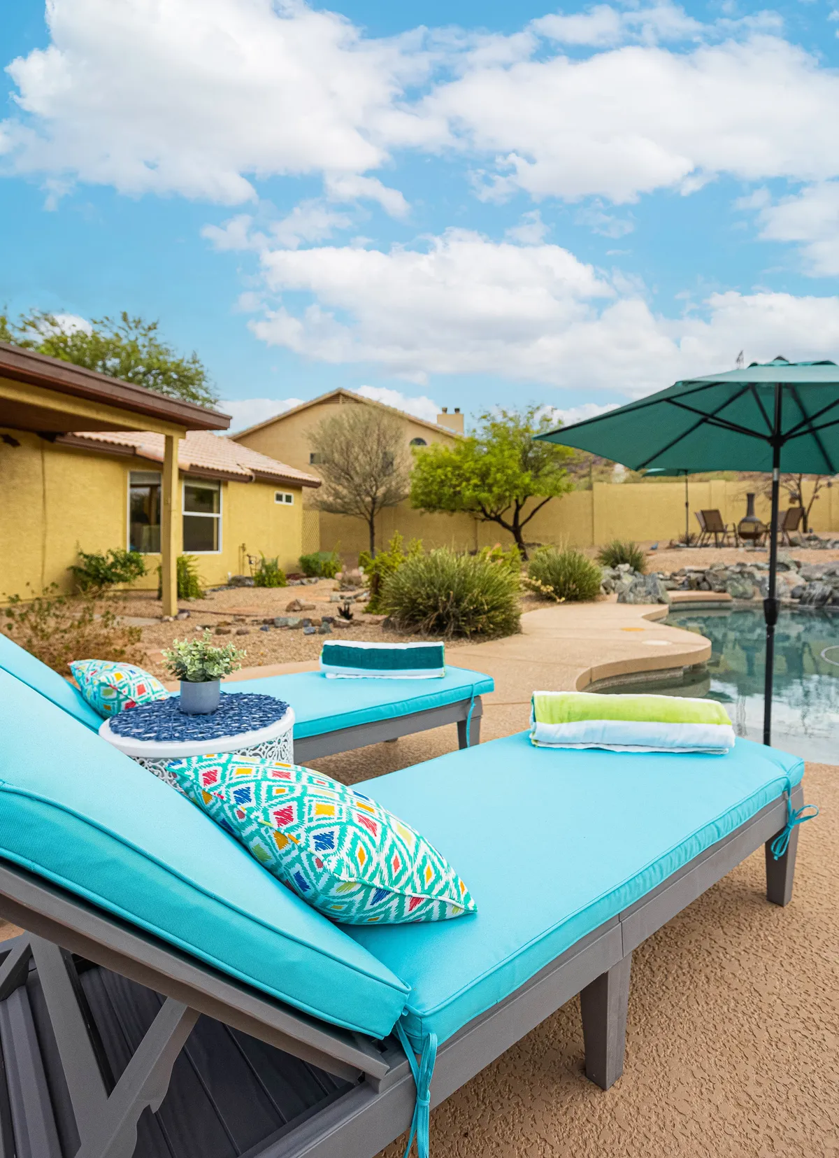 This image features a turquoise swimming pool surrounded by bright lounge chairs with colorful cushions and a small round table, perfect for relaxing or socializing. The sparkling water and inviting seating area evoke a sun-drenched, carefree atmosphere ideal for family fun and poolside gatherings.