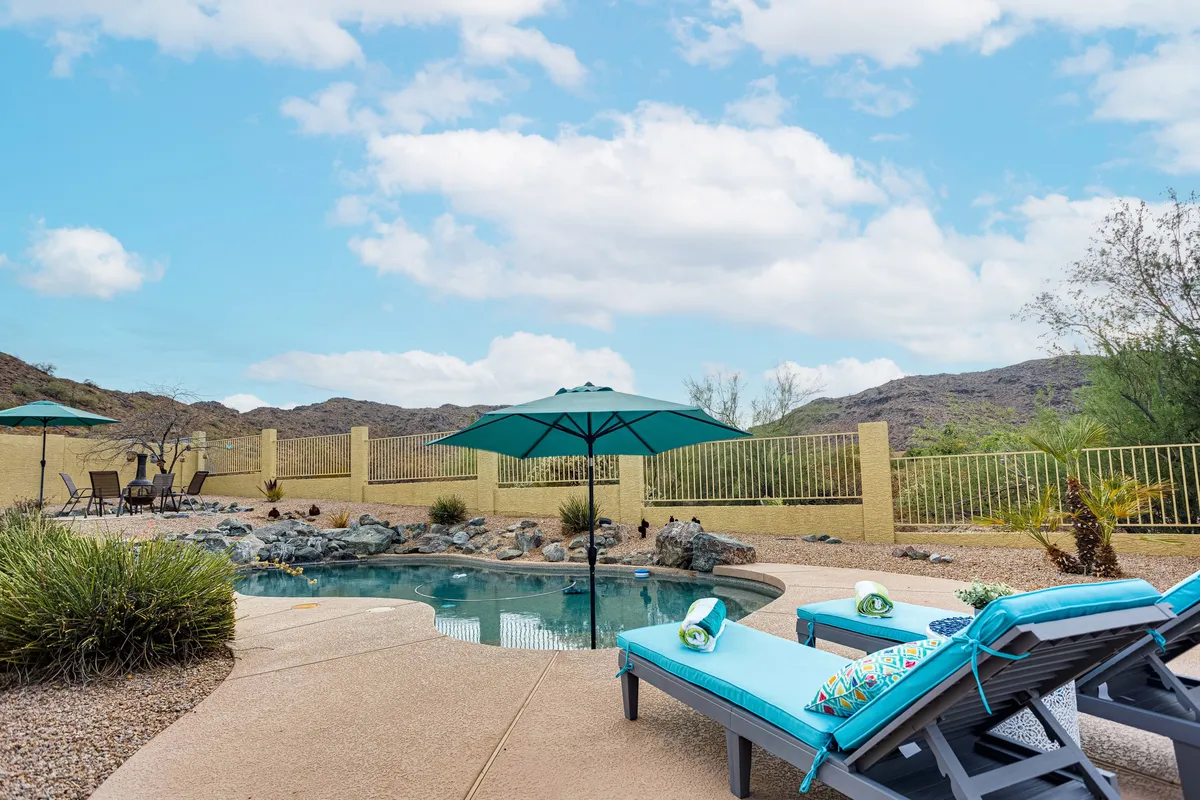 A tranquil backyard scene with a sparkling swimming pool, several lounge chairs, and an umbrella, all set against a stunning mountain backdrop. The setting sun casts a warm glow over the water and mountains.