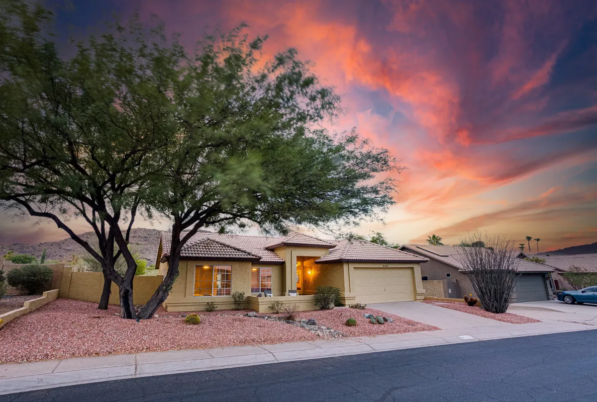 An exterior view of the house at sunset, with a warm glow from the windows and a large tree in the front yard, gives a welcoming, homey feel. The driveway is visible, suggesting ample space for both covered and additional parking.