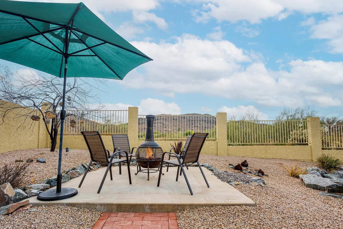 A cozy backyard patio featuring a fire pit surrounded by outdoor seating and shaded by an umbrella. The area is set for gatherings, with the glow of the fire pit suggesting warmth and comfort as the sun sets.