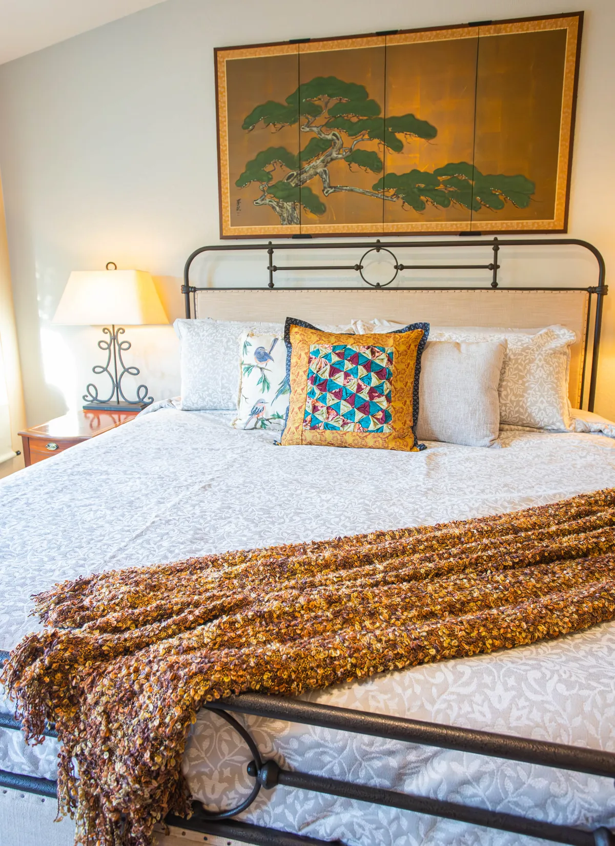 A cozy bedroom with a patterned bedspread, decorative throw blanket, colorful accent pillow, and calming wall art. Natural light streams in, creating a tranquil space ideal for restful sleep and quiet moments.