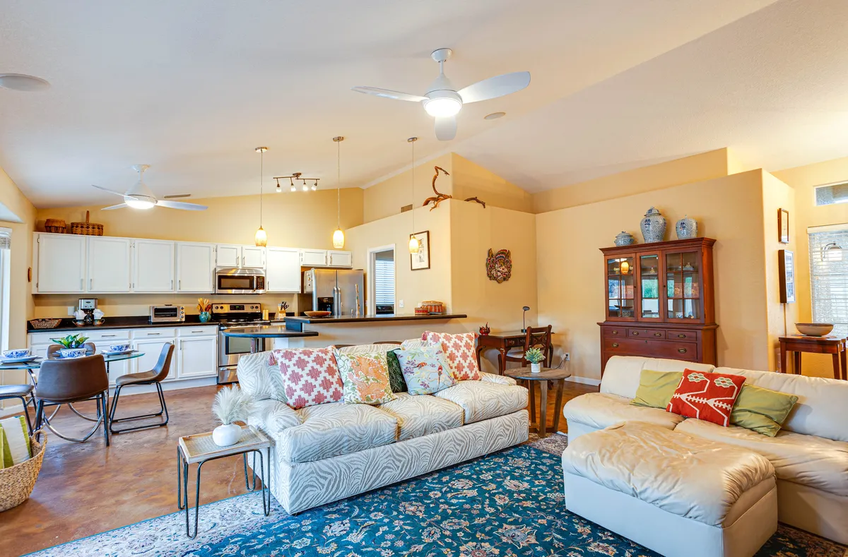 A homey open-concept interior displaying the sofa with decorative pillows, a dining table with chairs, and a modern kitchen area with visible appliances and a display cabinet. The seamless connection between the living, dining, and kitchen zones is evident, and the mood is inviting and family-oriented.